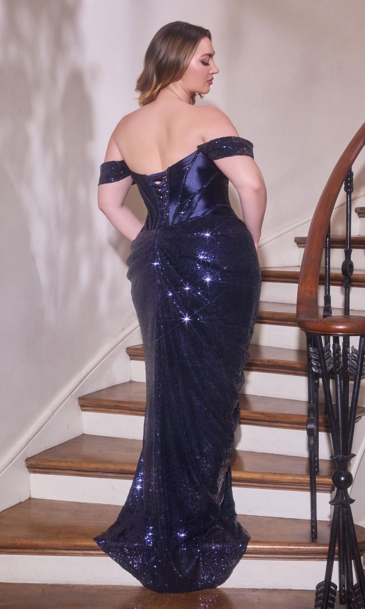 A woman stands on a wooden staircase, facing away, wearing the Ladivine CD422C long plus-size prom dress in dark blue with an off-shoulder neckline and sequined skirt.